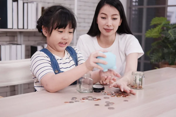 Smiling Asian little asian girl child is putting coins into piggy bank for saving money for the future with mother on wooden table. Child educational for homeschool concept.