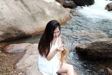 Beyaz elbiseli gülümseyen Asyalı kadın kayanın üzerinde oturuyor ve samrtphone ile online alışveriş için rahatlıyor. Tropikal ormanlarda açık hava ve akarsu teknolojisinin yaşam tarzı.