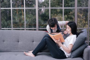 Gülümseyen Asyalı anne küçük Asyalı kıza kitap okumayı öğretiyor ve oturma odasında birlikte alfabeyi öğreniyoruz. Ev okulu ve eğitim konsepti