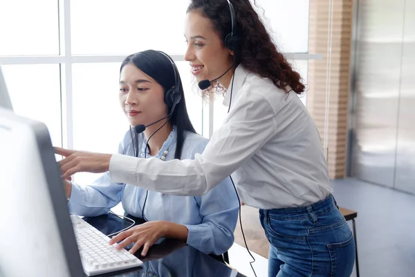 stock image Smling asian and caucasian women are colleagues and call center or secretary operator with headset and a microphone together. They are Consulting to solve problems about customers at office.