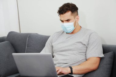 Maske takan genç Kafkas işadamı covid-19 virüsünün rahatlamasını ve aile evindeki oturma odasında sosyal medyada dizüstü bilgisayarla çalışmasını engelliyor. Teknoloji ve iş ev kavramı.
