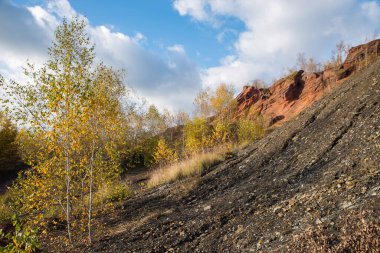 Sonbahar ağaçları ve mavi bulutlu gökyüzü olan kırmızı dağlar. Madenlerden kömür çıkardıktan sonra renkli atık yığınları.