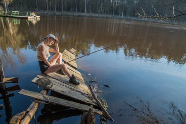 Ahşap bir iskelede elinde olta olan bir kız orman gölünde balık tutuyor..