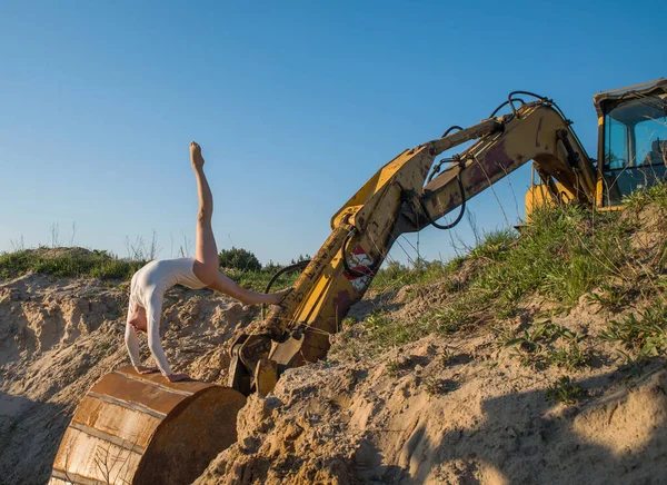 A girl in a white bodysuit in an open space among the sand near an old excavator.