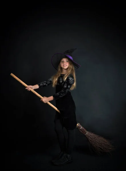 Egy kislány sötét háttérben boszorkányjelmezben, és egy széles karimájú kalap repül a seprűn Halloweenkor.. — Stock Fotó