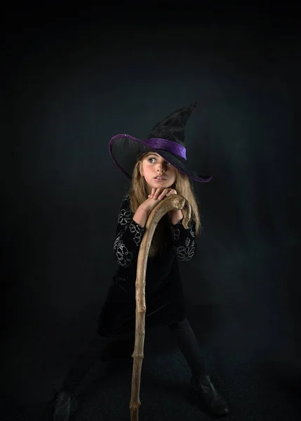 Una chica sobre un fondo oscuro en un disfraz de bruja y con un sombrero de ala ancha con un bastón para Halloween. —  Fotos de Stock