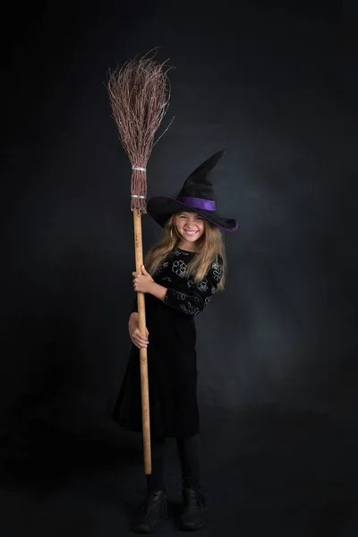 Cadılar Bayramı için süpürgeyle süslenmiş geniş kenarlı şapkalı siyah bir arka planda küçük bir kız.. — Stok fotoğraf