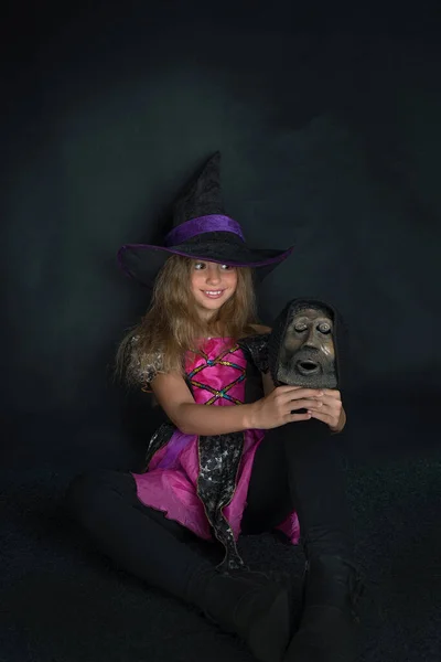 Una chica vestida de bruja y un sombrero de Halloween de ala ancha con la cabeza de un hombre barbudo en sus manos. —  Fotos de Stock
