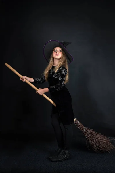 Een klein meisje op een donkere achtergrond in een heksenkostuum en een brede hoed vliegt op een bezem met Halloween. — Stockfoto
