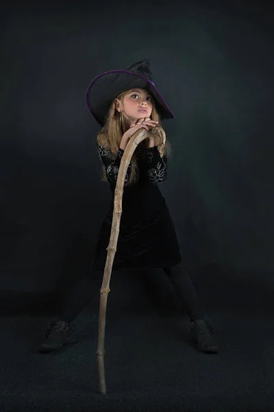 Egy lány sötét háttérben boszorkányjelmezben és széles karimájú kalapban, botokkal Halloweenkor.. — Stock Fotó