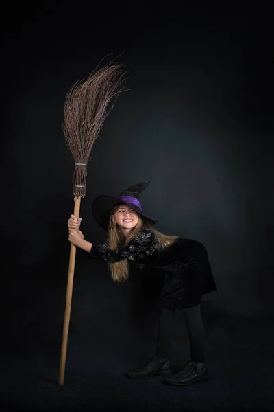 Egy Kislány Sötét Háttérben Boszorkányjelmezben Széles Karimájú Kalapban Seprűvel Halloweenkor — Stock Fotó