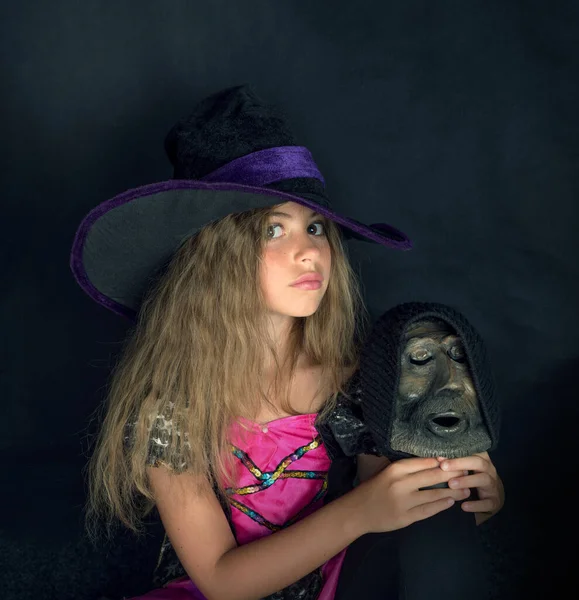 Una chica vestida de bruja y un sombrero de Halloween de ala ancha con la cabeza de un hombre barbudo en sus manos. —  Fotos de Stock