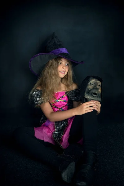 Una chica vestida de bruja y un sombrero de Halloween de ala ancha con la cabeza de un hombre barbudo en sus manos. —  Fotos de Stock