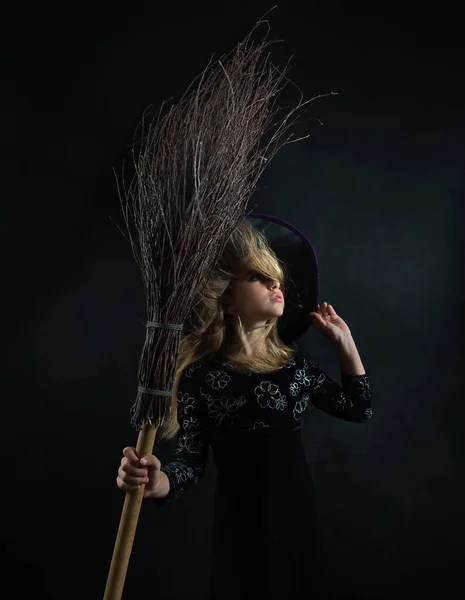 Klein meisje op een donkere achtergrond in een heksenkostuum en een brede hoed met een bezem voor Halloween. — Stockfoto