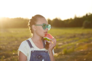 Bir kız gün batımında parkta lezzetli bir karpuz yiyor.