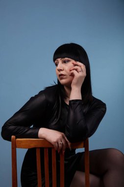 Siyah elbiseli, siyah saçlı güzel bir kız, esmer bir arka planda, eski bir tahta sandalyede erotik pozlar veriyor.