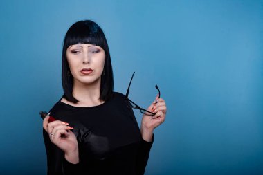 Koyu renk saçlı ve gözlüklü güzel bir kız mavi arka planda elektronik sigara içiyor.
