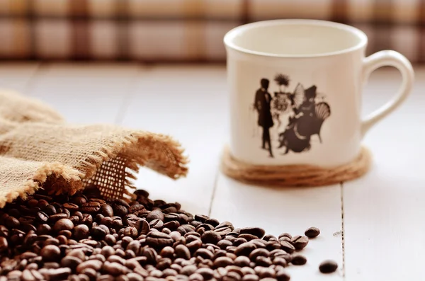 Spilled coffee beans from canvas sack and porcellaneous mug — Stock Photo, Image