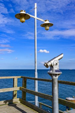 Almanya 'nın Kullanım Adası' ndaki Bansin 'deki rıhtımda madeni para yuvası ve fener bulunan bir teleskop. Deniz ve gökyüzü koyu mavi ve gün ışığında ışıldıyor..
