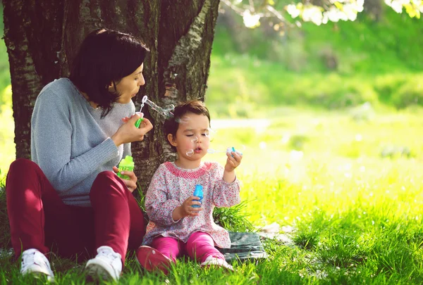 Happy mladá matka a její dcera foukání mýdlové bubliny v parku — Stock fotografie