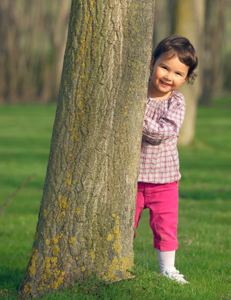 Menina bonita espreitando atrás de uma árvore — Fotografia de Stock