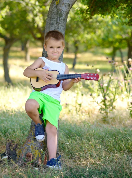 Menino bonito tocando guitarra perto da árvore de verão — Fotografia de Stock