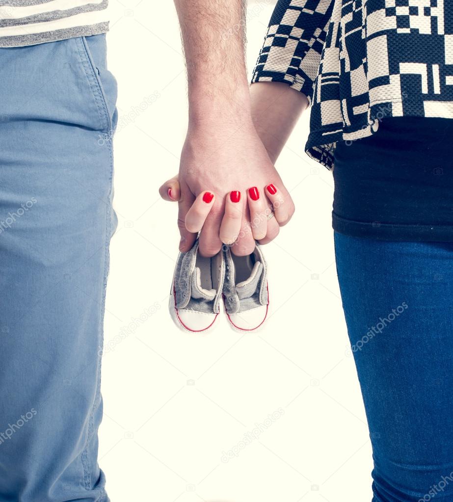Future parents holding hands and a pair of little shoes over white background
