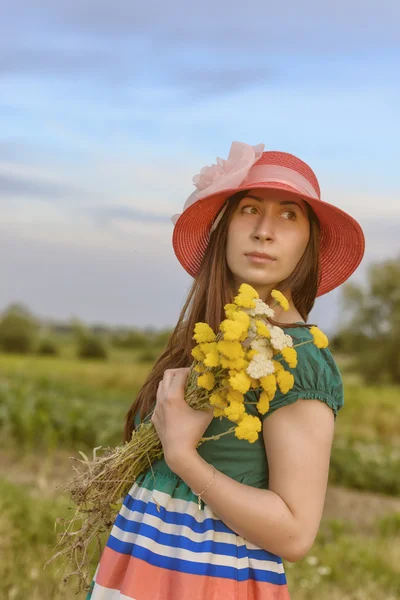 Porträt einer jungen Frau, die mit rotem Hut auf einem Feld steht und Blumen hält — Stockfoto