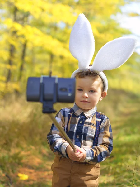 Funny little kid boy with Easter bunny ears taking a selfie with a selfie stick — Stock Photo, Image