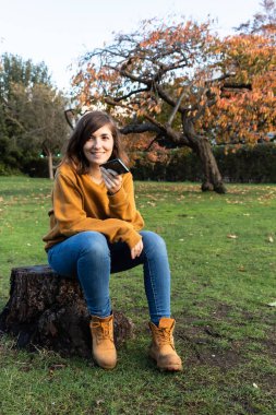 Kesilmiş antik bir ağacın dibinde oturan çekici genç bir kadın sonbahar sezonu boyunca Regents Park 'taki akıllı telefonuyla bir arkadaşına kayıt gönderiyor..