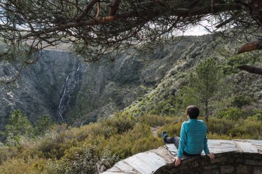 Genç ve çekici bir kadın, mavi ceketli, uzun bir yürüyüşten sonra kayalık bir duvarın tepesinde oturuyor ve önünde küçük bir şelaleye tanık oluyor..