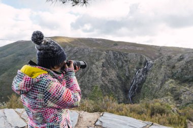 Renkli ceketli genç ve çekici bir kadın dijital kamerayla ormanda manzara fotoğrafları çekiyor..