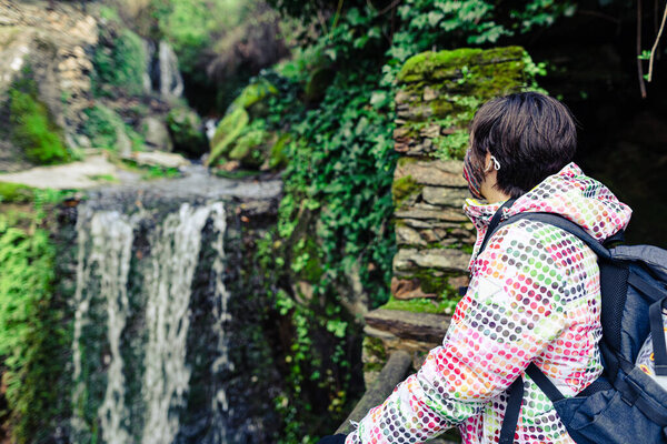 Young and attractive female with a colourful jacket standing and witnessing a small waterfall between houses in a small and ancient village in Spain.