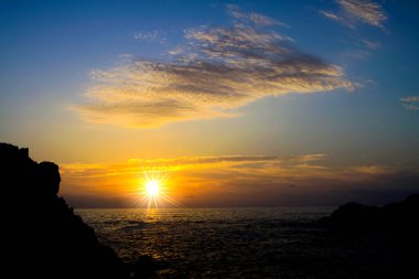 Atlantik Okyanusu üzerinde canlı sarı turuncu gün batımına bakın. Kayalardan ve bulutlardan oluşan siluet, güneş patlaması etkisi.