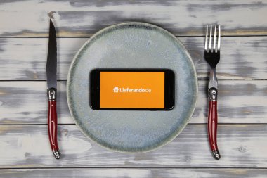 Viersen, Almanya - 9 Mayıs. 2020: Tabak ve çatal bıçak ile ahşap masa üzerinde yemek dağıtım servisi lieferando logosu ile cep telefonu ekranını kapat (merkeze odaklan)