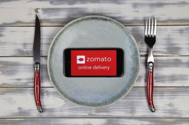 Viersen, Almanya - 9 Mayıs. 2020: Cep telefonu ekranının üzerinde Hint yemeği dağıtım servisi Zomato 'nun logosu bulunan tabaklar ve çatal bıçaklarla kapatılması (merkeze odaklanma))