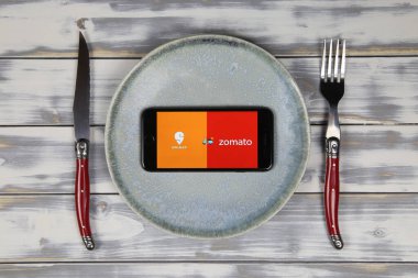 Viersen, Almanya - 9 Mayıs. 2020: Cep telefonu ekranının üzerinde Hint yemeği dağıtım servisi logosu olan ahşap masadaki tabak ve çatal bıçaklı Swiggy Zomato 'yu kapat (merkeze odaklan)