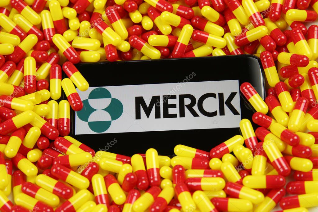 Viersen, Germany - April 9. 2020: Close up of mobile phone screen with logo lettering of Merck pharmaceutical company on pile yellow red drug capsules (focus on center of lettering)