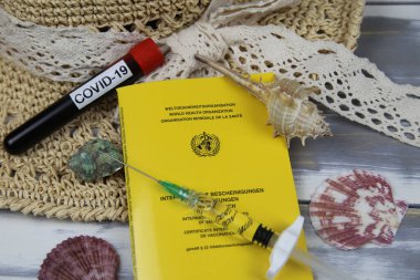 Viersen, Almanya - 1 Mart. 2021: Ahşap masa üzerinde covid-19 kan örneği şişesi, şırınga, güneş şapkası ve deniz kabuğu ile aşılama uluslararası sertifikalarının kapatılması (ortada WHO logosuna odaklanın)