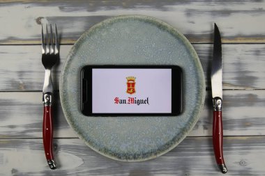 Viersen, Almanya - 9 Mayıs. 2021: Tabak ve ahşap masadaki cep telefonunun kapatılması, San Miguel şirketinin yiyecek ve içecek şirketinin logosu olan çatal bıçak takımı (merkeze odaklan)