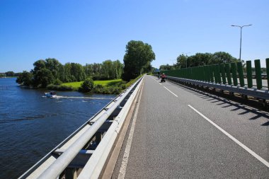 Mavi yaz gökyüzüne karşı yeşil manzaralı güzel göl boyunca (noorderplas) köprüdeki bisiklet pistine bakın - Roermond, Hollanda