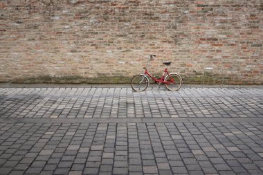 Eski kırmızı bir bisiklet kaldırım taşlarının önündeki tuğladan duvara yaslanıyor, yazı için yeri var.