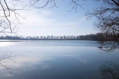 Kışın, çeşitli ağaçların çıplak dalları Bavyera 'da gökyüzü olan bir gölün etrafında bir çerçeve oluşturur.