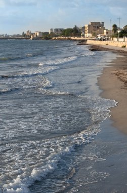 Sahilde, Mazara del Vallo 'nun önünde, ön planda. Arka planda Akdeniz 'deki şehir var. Güneş batıyor. Dalgalar kıyıya çarpıyor..