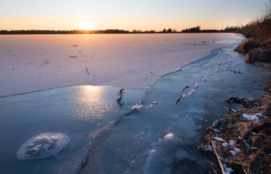 Kışın manzara çekimi. Ufukta donmuş bir göl. Güneş doğuyor. Tahta ve sopa parçaları buzun içinde donmuş. Sahil kenarda..