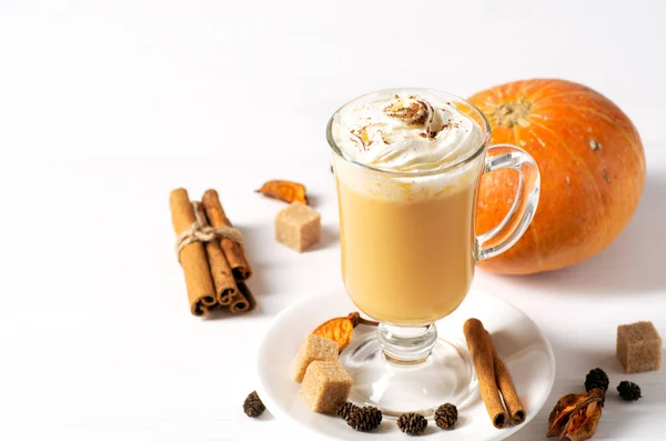 A beautiful glass with pumpkin latte and whipped cream stands on a white background, next to it lies pumpkin and cinnamon. Horizontal orientation, copy space.