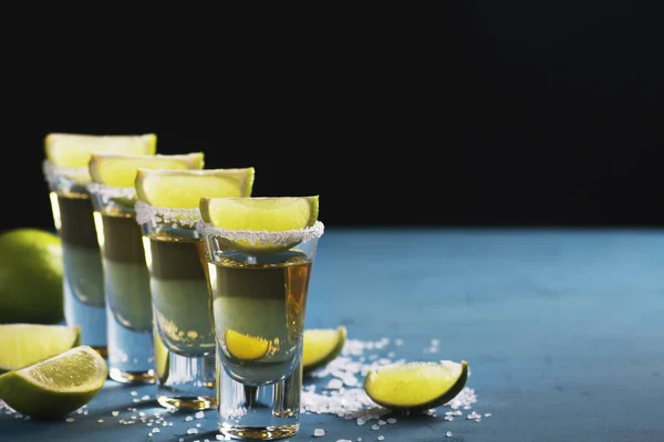 Shots with gold Mexican tequila, decorated with salt and lime, stand against a dark background, copy space, horizontal orientation.