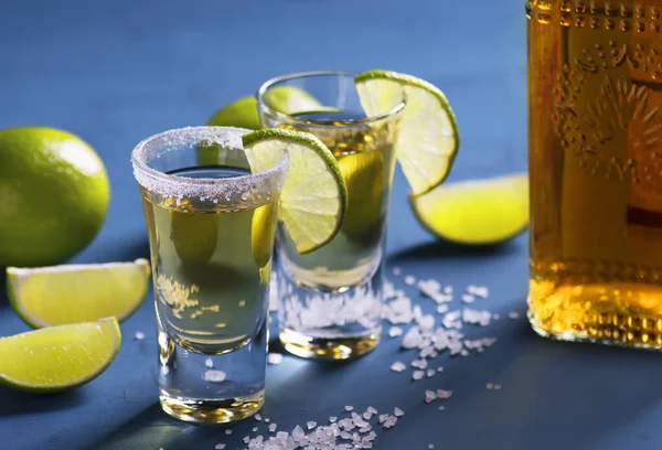 Shots with Mexican tequila, limes, salt, bottle with golden tequila stand on a blue background, selective focus, close-up, horizontal orientation.