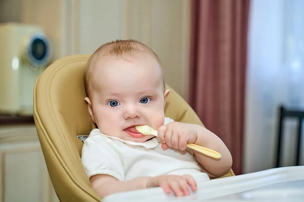 Un niño pequeño con ojos azules sostiene una cuchara sentada en una silla alta. — Foto de Stock