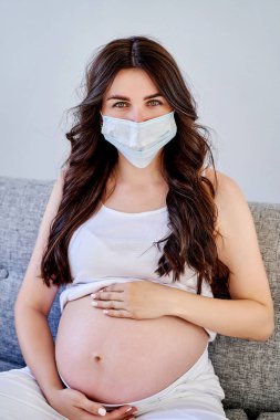 Tıbbi maskeli ve beyaz tişörtlü güzel hamile kadın pencerenin yanında karantinaya alındı. Coronavirus, virüs.